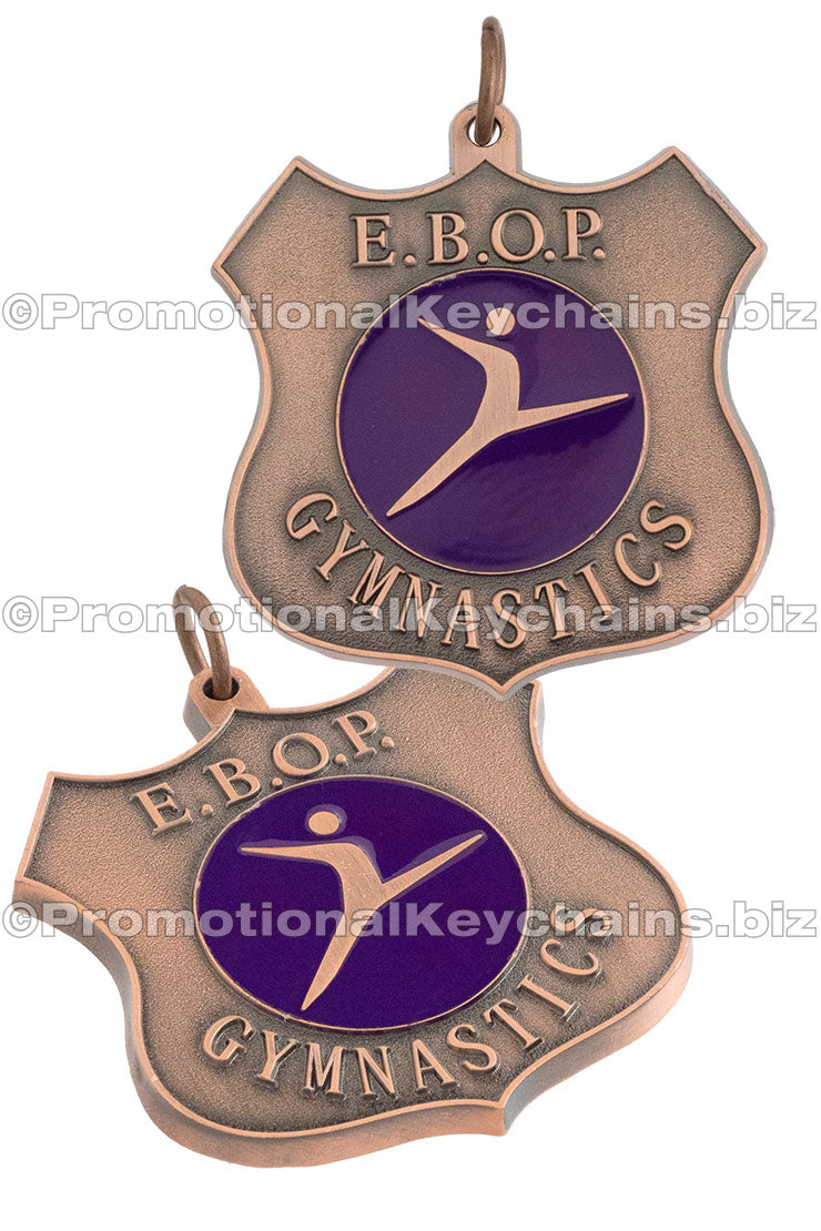Two angles of an antiqued copper medal, shaped like a shield with a purple enamel emblem in the center and the text "E.B.O.P. GYMNASTICS" around the outside.