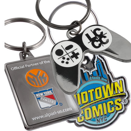 An assortment of three polished black metal keychains, featuring a game controller design, a rectangular design featuring New York sports team logos, and and a design with the Midtown Comics logo.