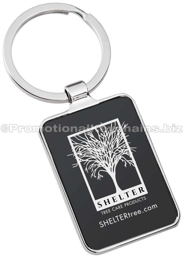 A polished metal keychain featuring a black rectangular background with a tree logo and the text "SHELTER Tree Care Products" engraved in the center.