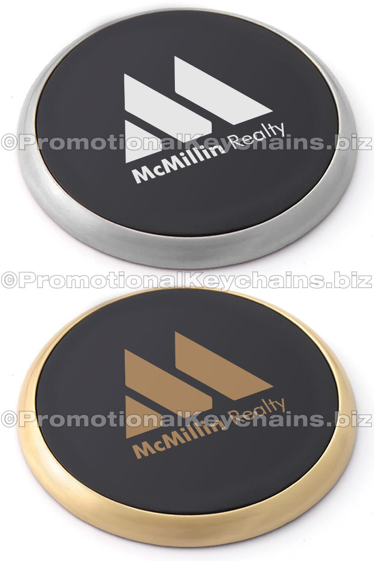There are two images of the same coaster design in different color platings. The top is in nickel and the bottom is in brass. They both have a black leather center with an imprint of the McMillin Realty logo.