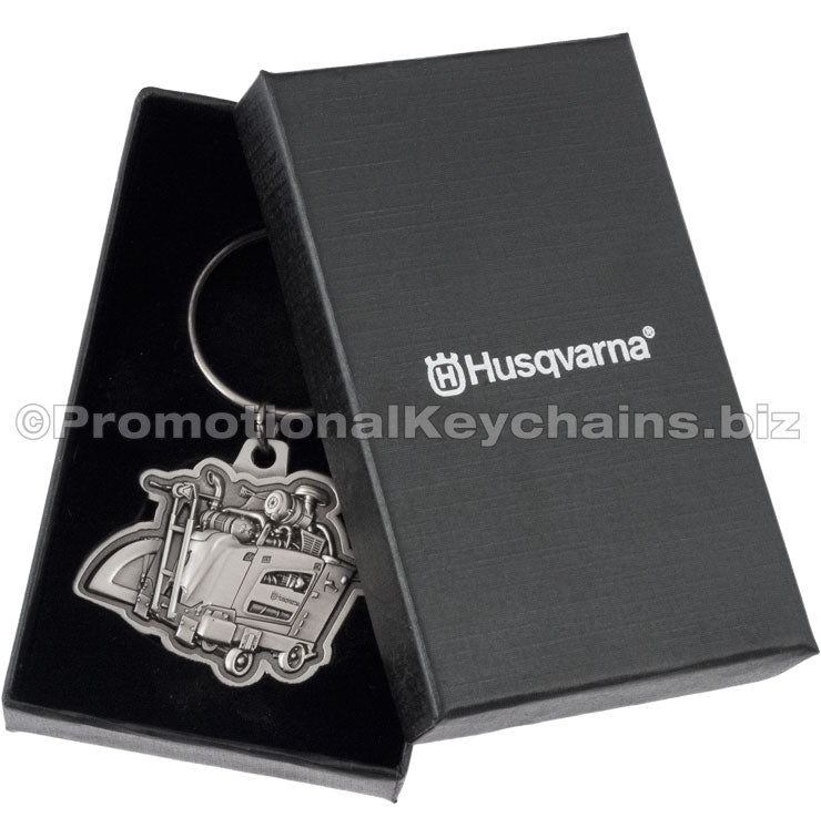 An antiqued nickel keychain featuring a detailed, 3-dimensional flat saw design placed into a black gift box. The lid of the box is visible to the side and has a custom silver imprint on the center.