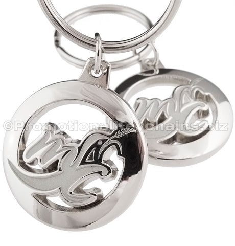 Two angles of the same circular polished nickel keychain, featuring a stylized "MC" logo cutout.