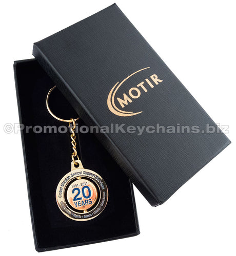 A polished gold spinning keychain commemorating a 20 year anniversary in blue enamel color, presented in a black gift box with a custom gold imprint on the lid.