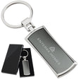 There are two images. On the left, a polished metal keychain with a black inlay displayed in a black gift box. On the right is the same keychain with a logo clearly engraved in the center.
