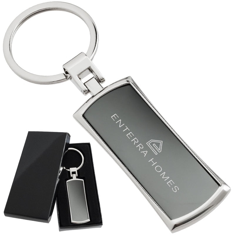 There are two images. On the left, a polished metal keychain with a black inlay displayed in a black gift box. On the right is the same keychain with a logo clearly engraved in the center.