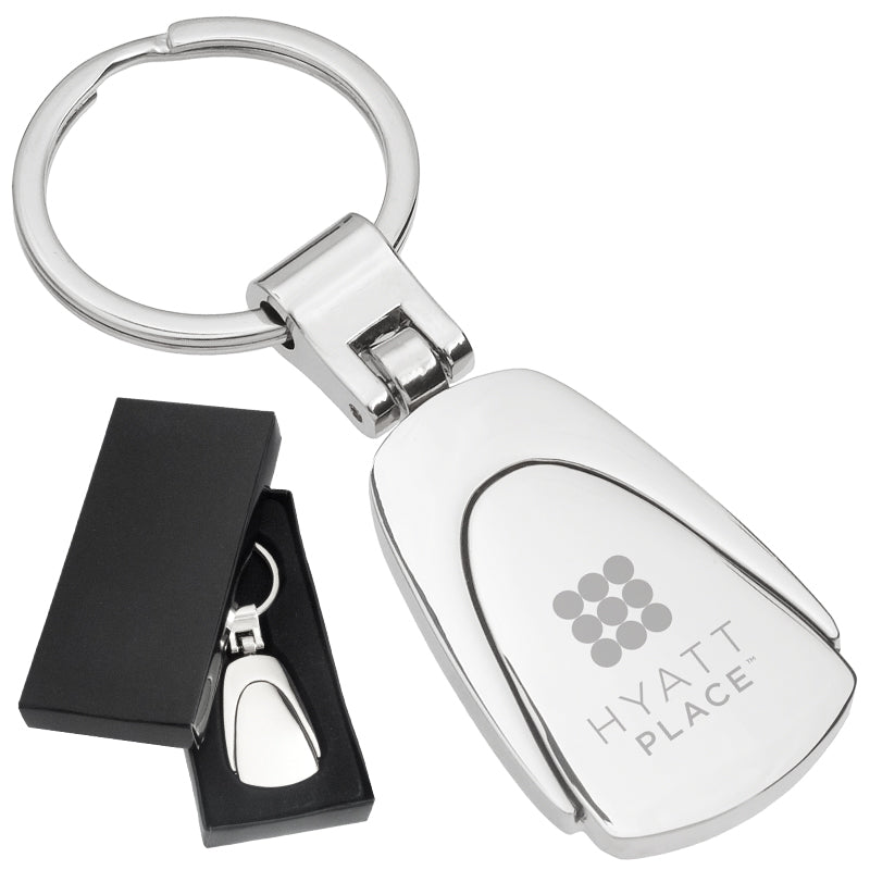 There are two images. On the left, a polished metal fob keychain displayed in a black gift box. On the right is the same keychain with a logo clearly engraved in the center.