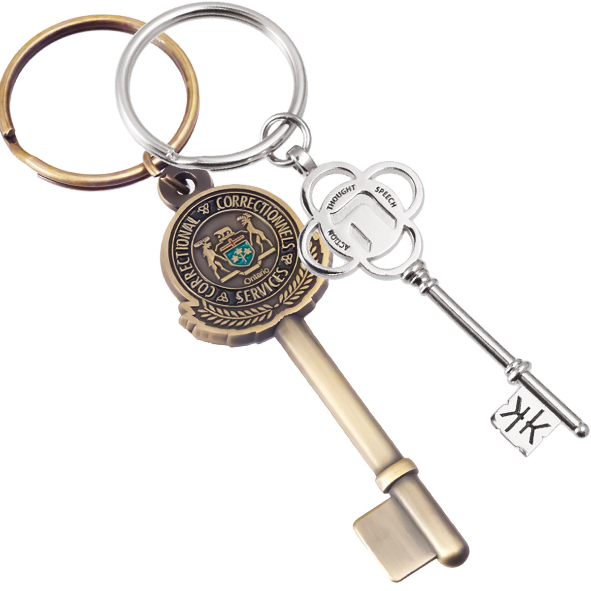 A photo of two different keychains. On the left, an antiqued brass keychain shaped like a key with a circular Ontario Correctional Services emblem embossed on the bow. On the right, a polished nickel keychain designed as a decorative key with a four-petal flower, engraved with the words "Thought," "Speech," and "Action." The tip of the key also has a debossed design.