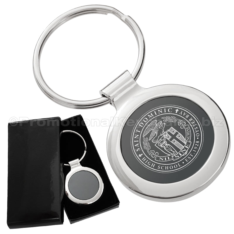 There are two images. On the left, a circular polished metal keychain featuring a black inlay displayed in a black gift box. On the right is the same keychain with a logo clearly engraved in the center.