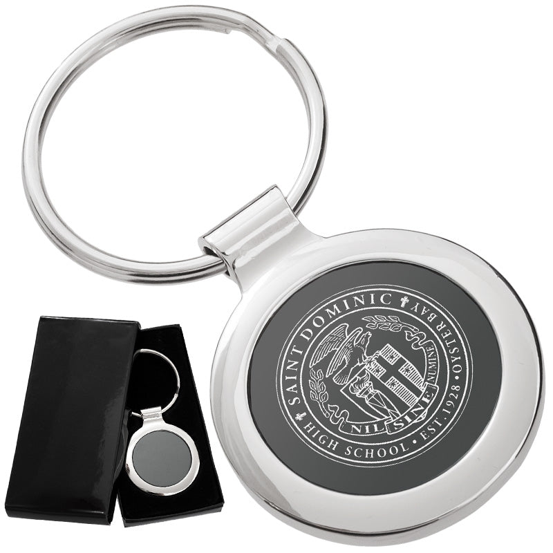 There are two images. On the left, a circular polished metal keychain featuring a black inlay displayed in a black gift box. On the right is the same keychain with a logo clearly engraved in the center.