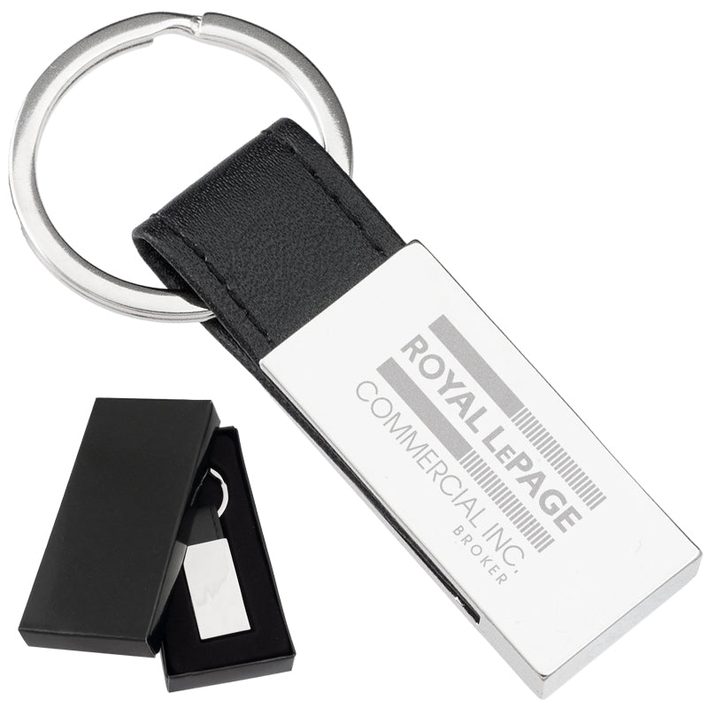 There are two images. On the left, a polished rectangular keychain with a black leather strap displayed in a black gift box. On the right is the same keychain with a logo clearly engraved in the center.