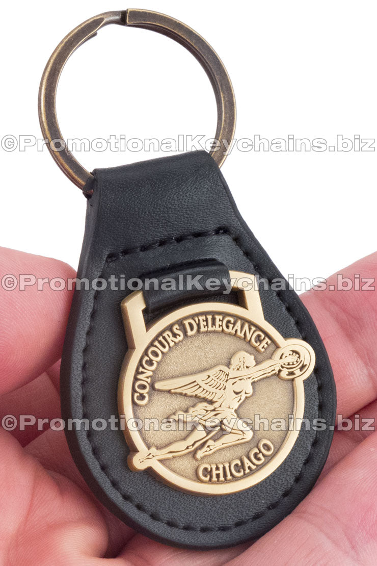 A black leather fob keychain featuring the "Concours d'Elegance" emblem embossed on a circular antiqued brass medallion.