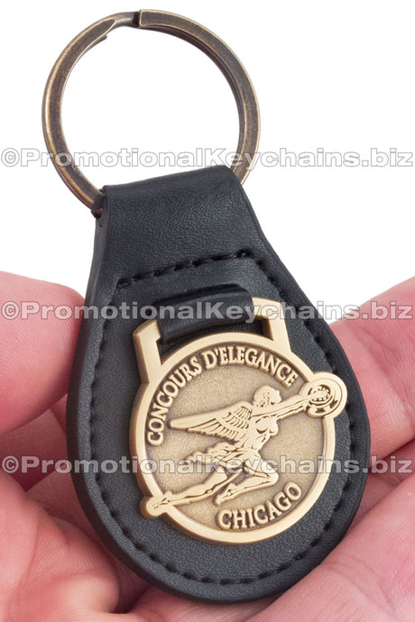 A black leather fob keychain featuring the "Concours d'Elegance" emblem embossed on a circular antiqued brass medallion.