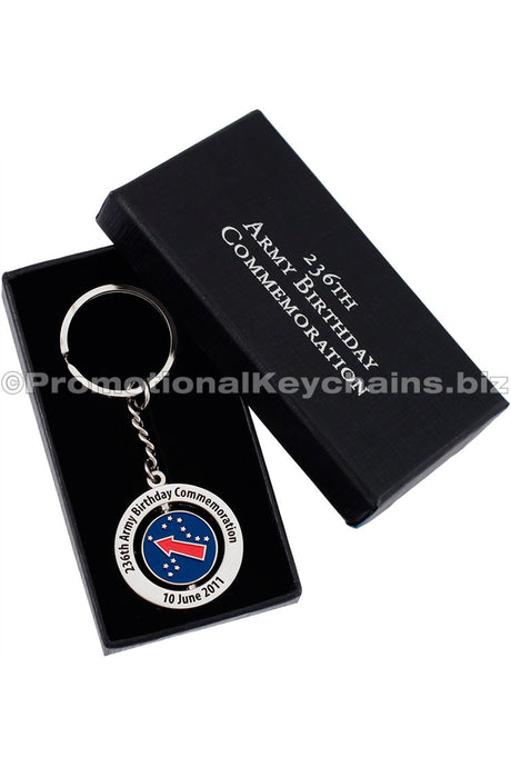 A polished nickel spinning keychain. with here is black text around the outer ring and a center design that has a blue background and red arrow with stars. It is presented in a black gift box, and there is a custom silver imprint on the lid.