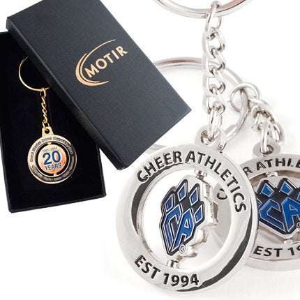 There are two product images. On the left, a polished gold spinning keychain commemorating a 20 year anniversary in blue enamel color, presented in a black gift box with a custom gold imprint on the lid. On the right, two different angles of the same polished nickel spinning keychain featuring a "CA" logo shaped like a paw in blue enamel in the center and black text around the outer ring.