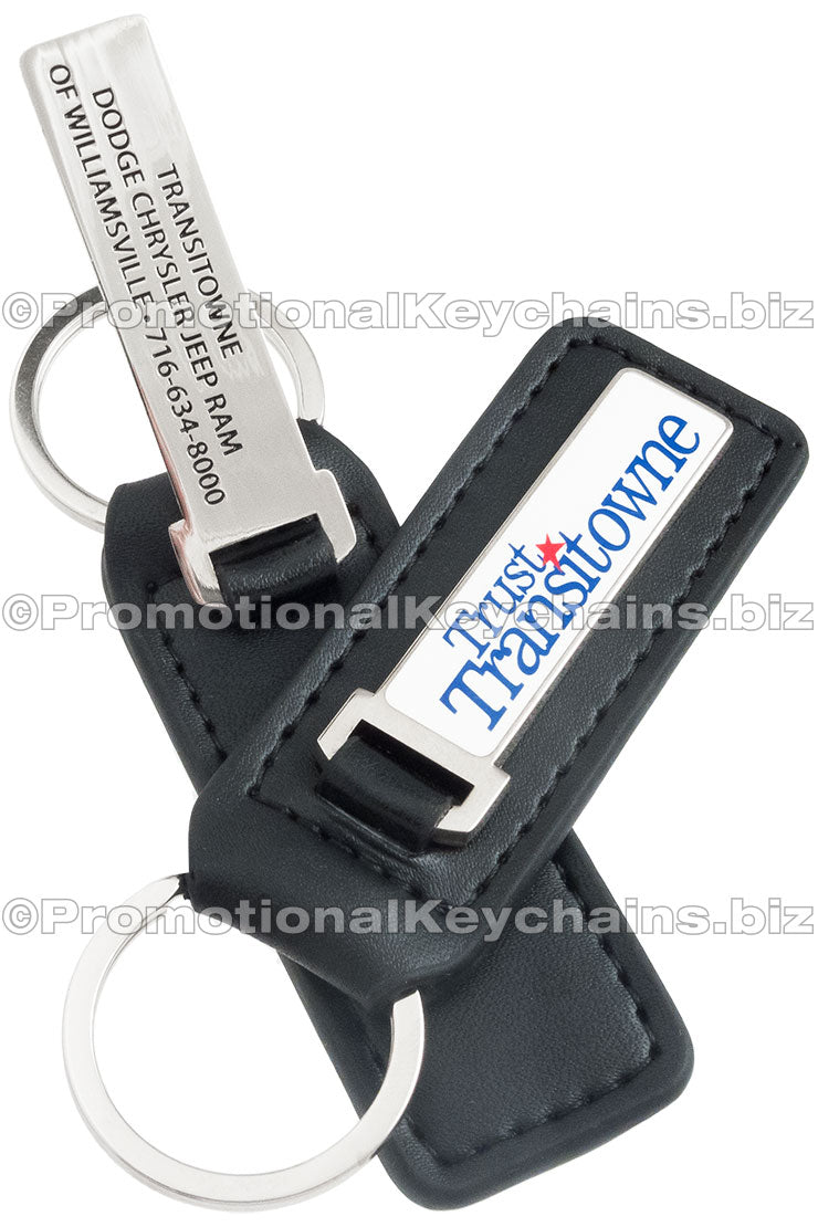 Two sides of the same fob keychain, featuring a sleek black leather design with an attached rectangular metal component, one side with the blue and red logo of Trust Transitowne, and the other side with the business' contact information.