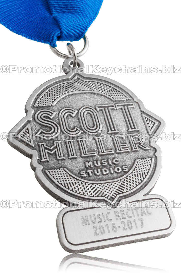 An antiqued nickel medal with blue ribbon, embossed with "Scott Miller Music Studios" in the center and "Music Recital 2016-2017" engraved on the bottom section.