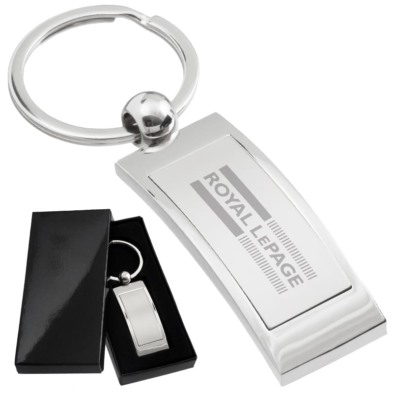 There are two images. On the left, a polished metal keychain, rectangular in shape, displayed in a black gift box. On the right is the same keychain with a logo clearly engraved in the center.