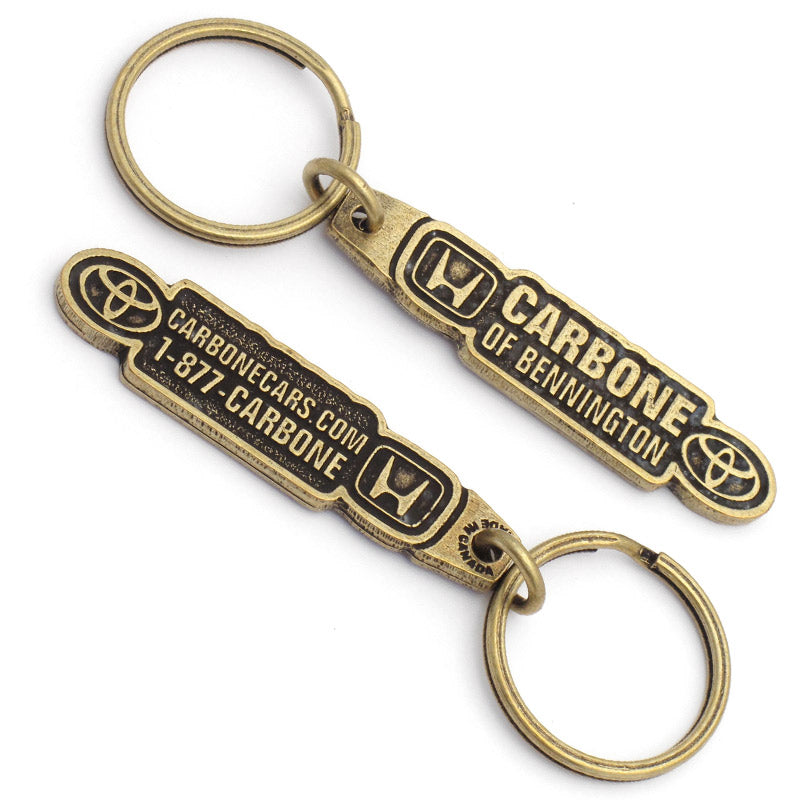 The front and back of the same antiqued brass keychain, featuring "Carbone of Bennington" with the Honda logo on the left and the Toyota logo on the right. The back has a phone number and a website.