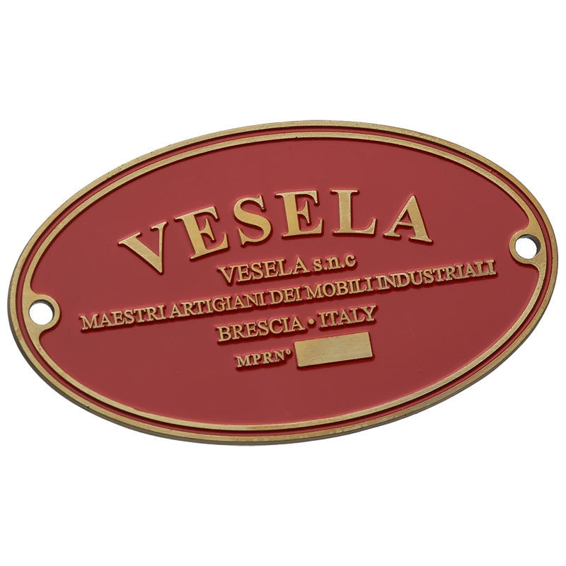 An oval plate, plated in antiqued gold with a red enamel background, showcasing the logo for "Vesela", an artisan furniture brand from Brescia, Italy. There are rivet holes on each end.
