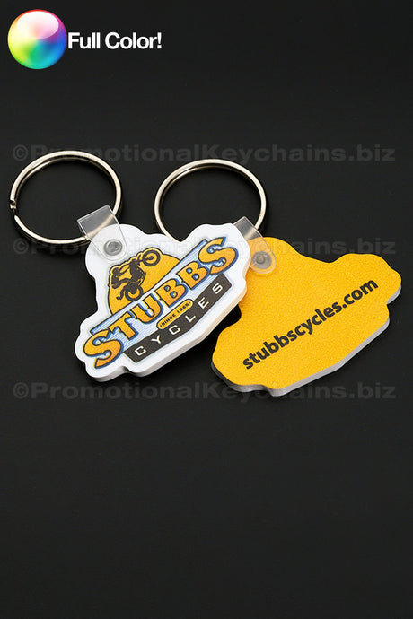 A white and a bright yellow vinyl keychain in the shape of the Stubbs logo with a printing of the logo on the front in full color against a dark background..