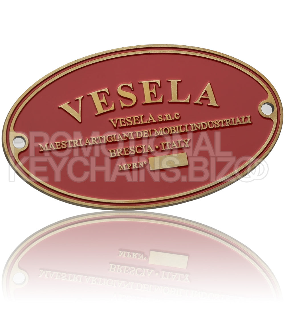 An oval plate, plated in antiqued gold with a red enamel background, showcasing the logo for "Vesela", an artisan furniture brand from Brescia, Italy. There are rivet holes on each end.