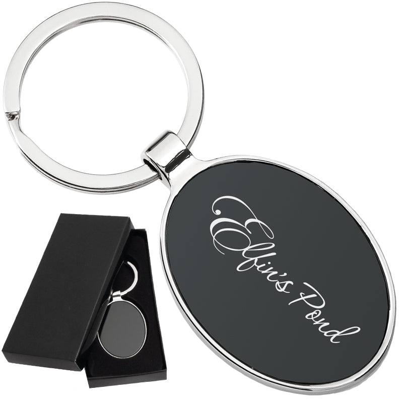There are two images. On the left, a polished metal keychain with an oval black center displayed in a black gift box. On the right is the same keychain with a logo clearly engraved in the center.