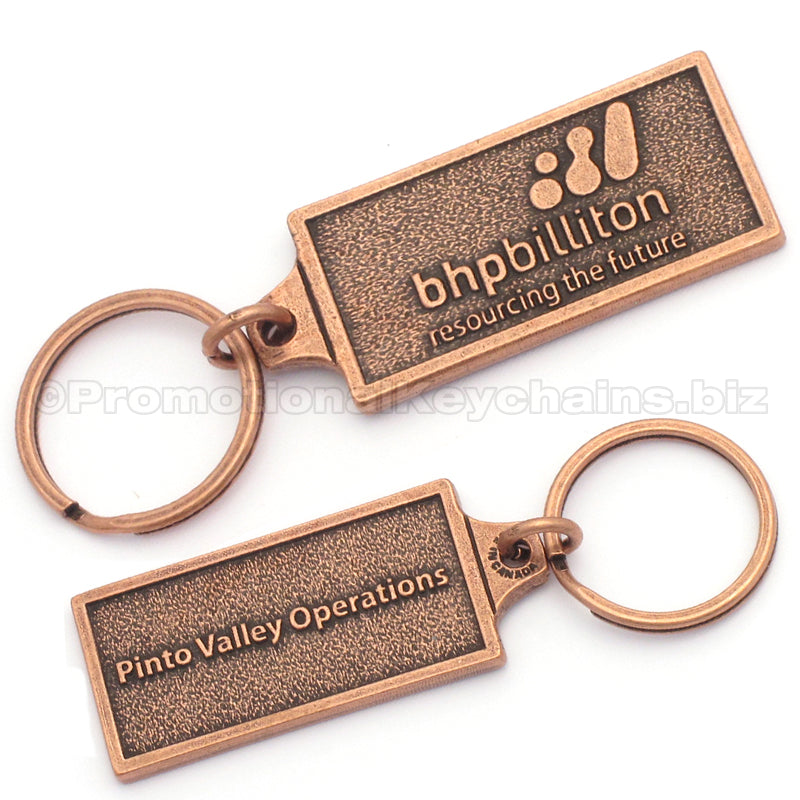 The back and front of a rectangular antique copper keychain. The front has the logo for BHPBilliton embossed, and the back has the text "Pinto Valley Operations" embossed.