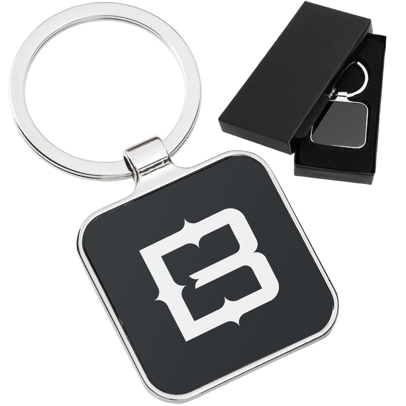 There are two images. On the right, a polished metal keychain featuring a square black background displayed in a black gift box. On the left is the same keychain with a logo clearly engraved in the center.