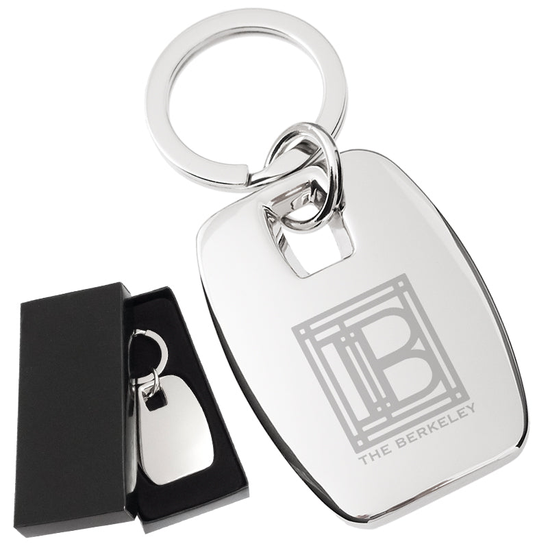There are two images. On the left, a polished metal rectangular keychain displayed in a black gift box. On the right is the same keychain with a logo clearly engraved in the center.