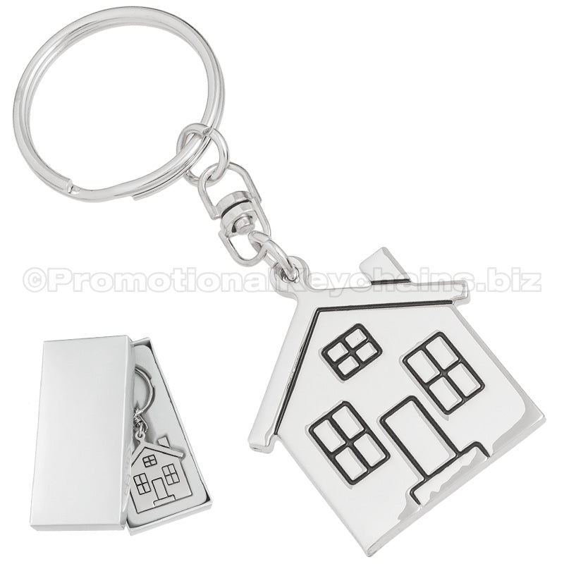There are two images. On the left, a polished metal house-shaped keychain displayed in a silver gift box. On the right is the same keychain, zoomed in for better detail.