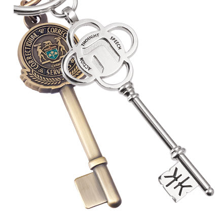 A photo of two different keychains. On the left, an antiqued brass keychain shaped like a key with a circular Ontario Correctional Services emblem embossed on the bow. On the right, a polished nickel keychain designed as a decorative key with a four-petal flower, engraved with the words "Thought," "Speech," and "Action." The tip of the key also has a debossed design.