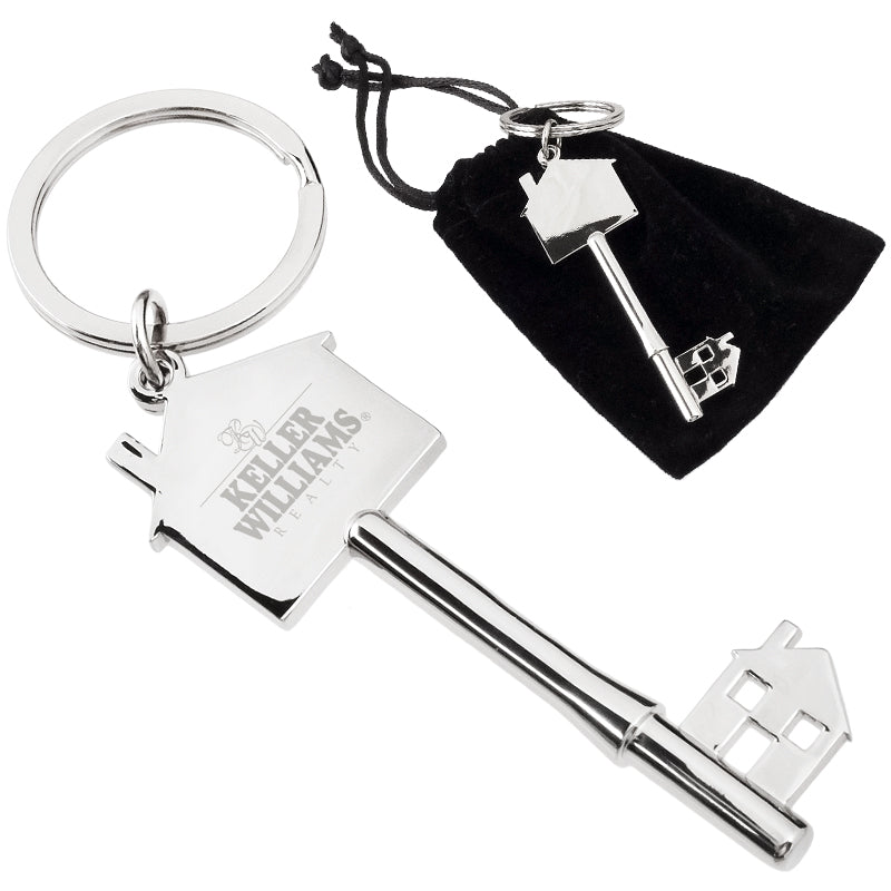 There are two images. On the right, a polished key-shaped keychain featuring a house silhouette, resting on top of a black velvet drawstring bag. On the left is the same keychain with a logo clearly engraved in the center.