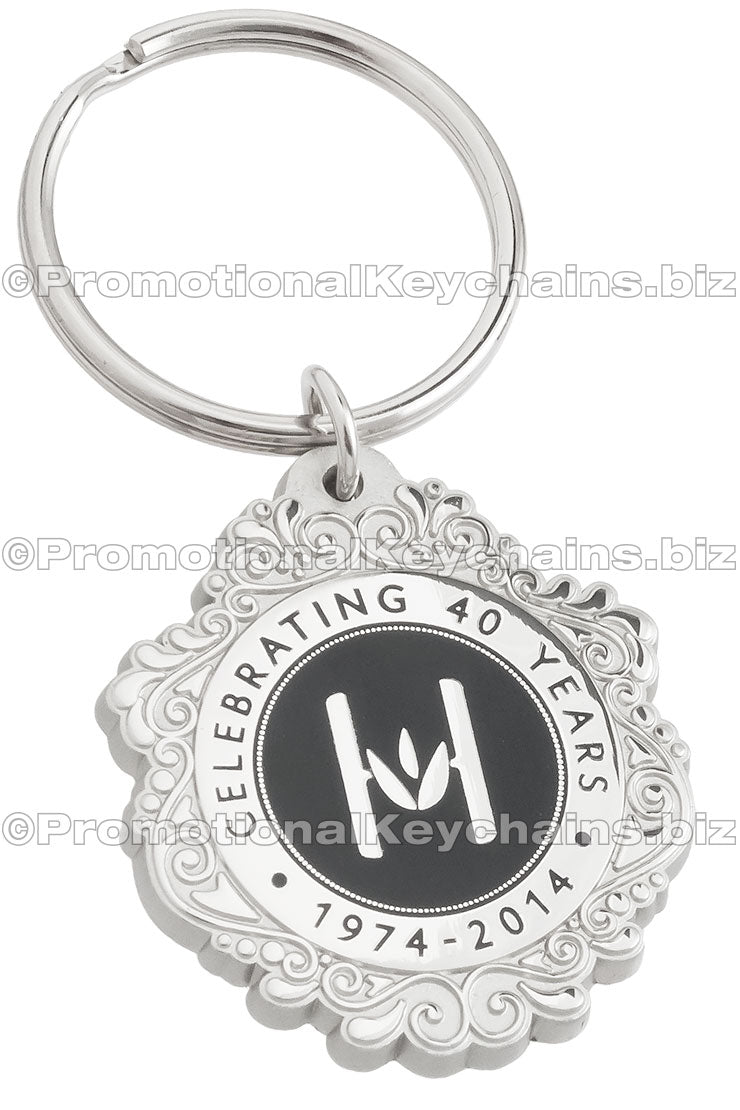 A circular polished nickel keychain with a black hard enamel center, featuring an "H" logo and an ornate border design.