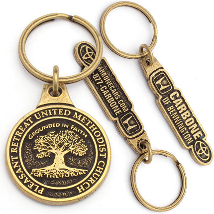 There are two different antiqued brass keychains pictured. The first is a circular design for the Pleasant Retreat United Methodist Church with a tree embossed in the center. The second design is shown with a photo of both sides of the keychain. The front has "Carbone of Bennington" with the Honda logo on the left and the Toyota logo on the right. The back has a phone number and a website.