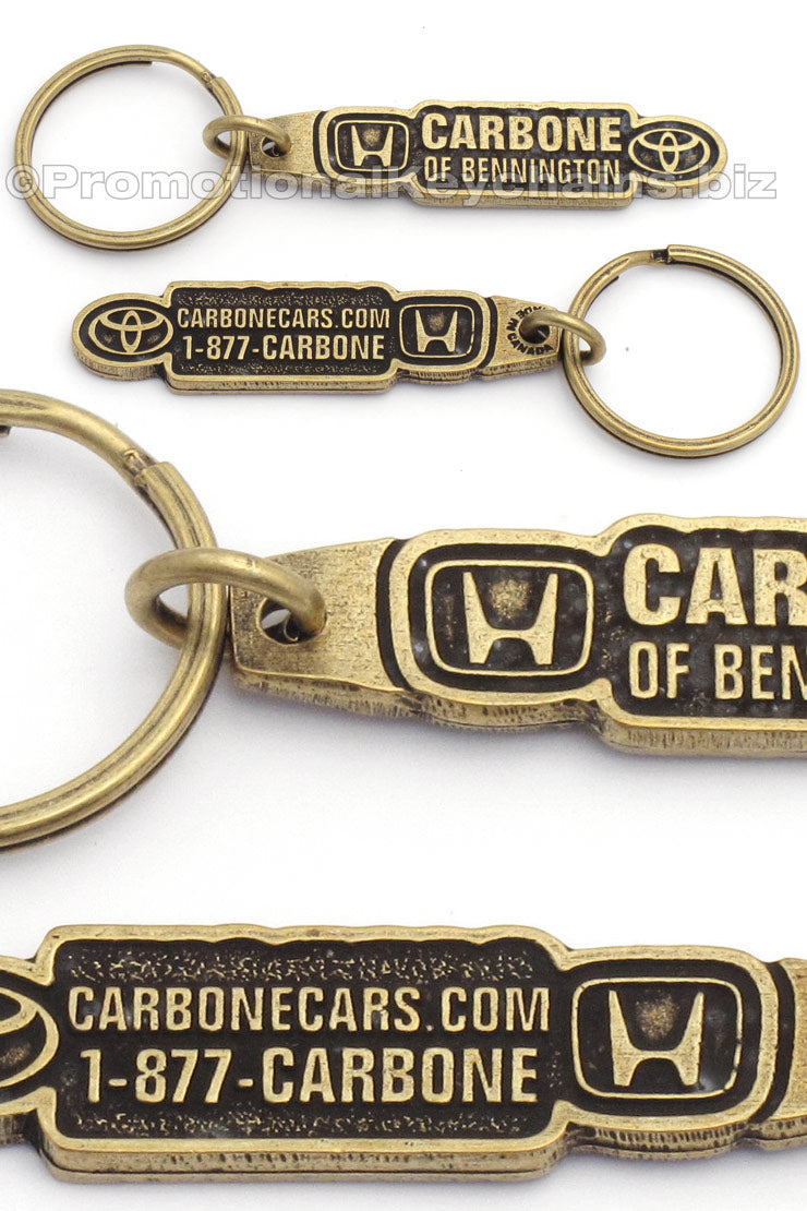 Multiple photos of the same antiqued brass keychain, featuring "Carbone of Bennington" with the Honda logo on the left and the Toyota logo on the right. The back has a phone number and a website.