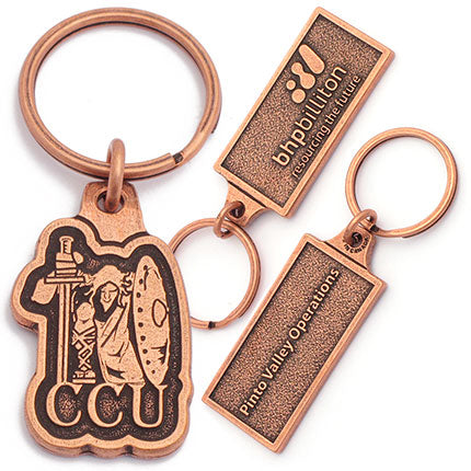 A photo of two different antiqued copper keychain designs. On the left design, there's a depiction of a figure holding a shield and sword on top of the text "CCU'. On the right, the front and back of the same rectangular antique copper keychain is shown. The front has the logo for BHPBilliton embossed, and the back has the text "Pinto Valley Operations" embossed.