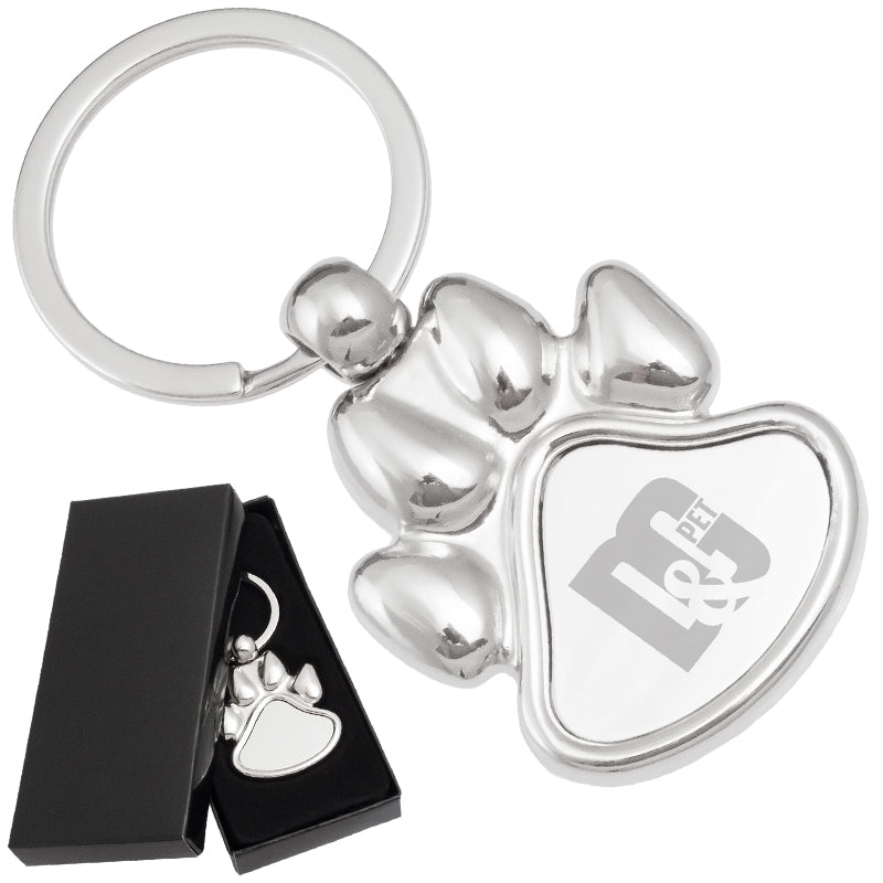 There are two images. On the left, a polished metal paw-shaped keychain displayed in a black gift box. On the right is the same keychain with a logo clearly engraved in the center.