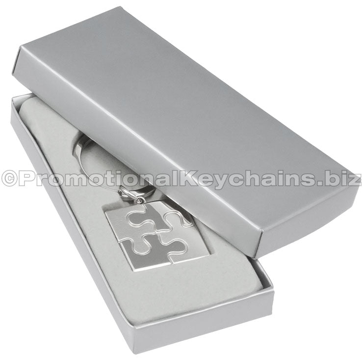 The font of a square keychain with a puzzle piece motif molded within the square. It is displayed in a silver gift box.
