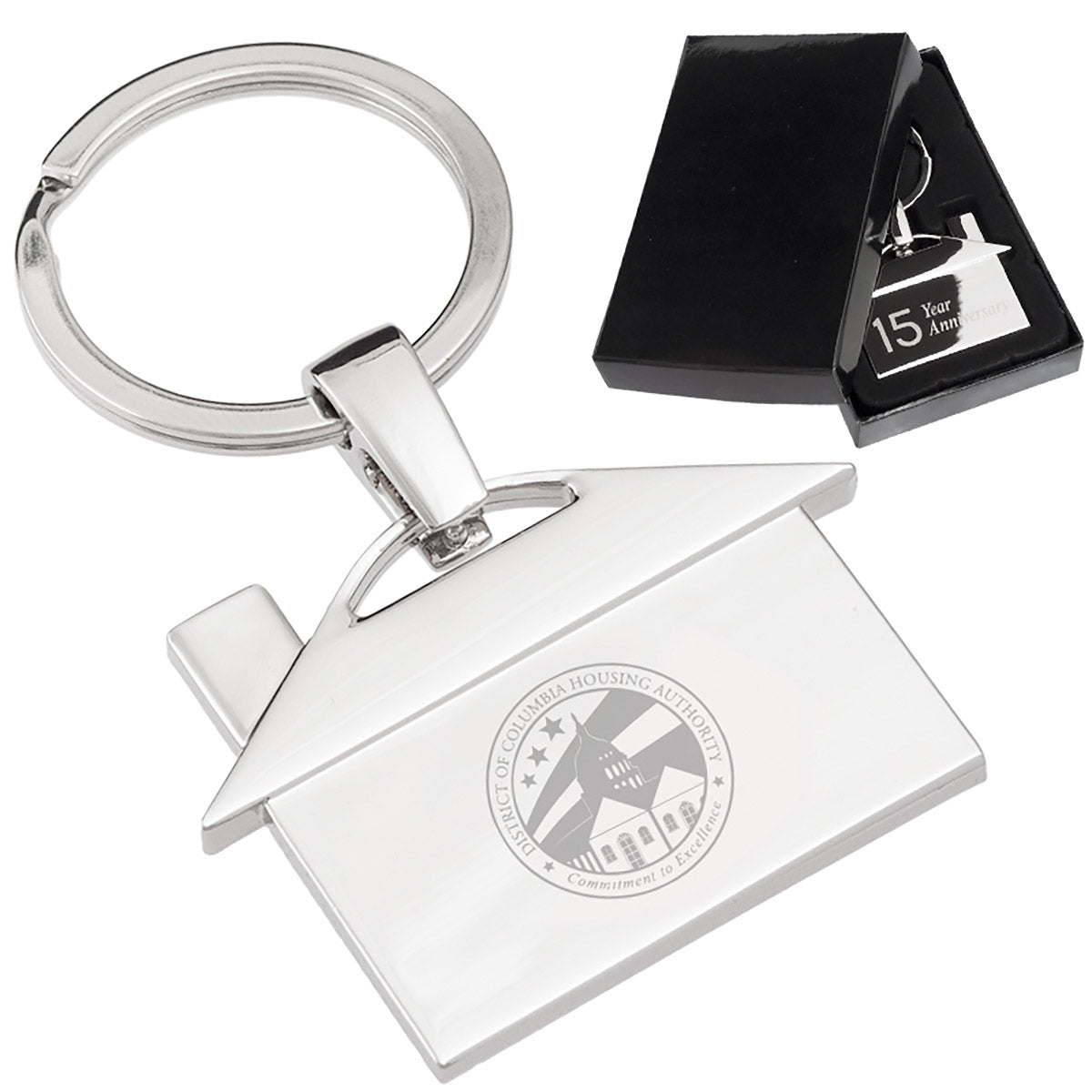 There are two images. On the right, a polished metal house-shaped keychain featuring a '15 Year Anniversary" engraving, displayed in a black gift box. On the left is the same keychain with a different logo clearly engraved in the center.
