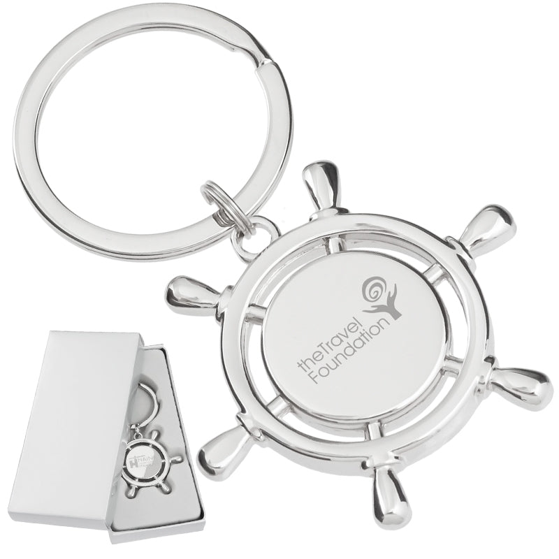 There are two images. On the left, a polished metal keychain that is shaped like a ship's wheel and laser engraved in the center. It is displayed in a silver gift box. On the right is the same keychain with a logo clearly engraved in the center.