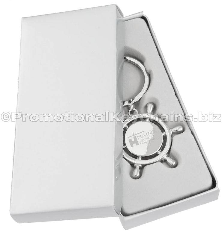A polished metal keychain that is shaped like a ship's wheel and laser engraved in the center. It is displayed in a silver gift box.