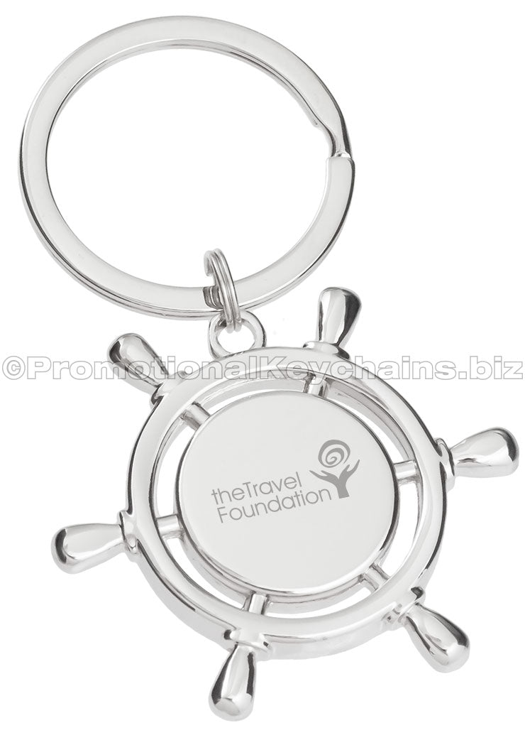 A polished metal keychain that is shaped like a ship's wheel, engraved with the logo for The Travel Foundation.