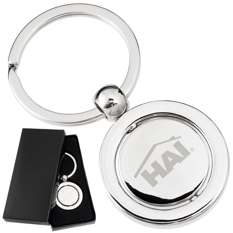 There are two images. On the left, a polished keychain featuring a round spinning disc in the center, displayed in a black gift box. On the right is the same keychain with a logo clearly engraved in the center.