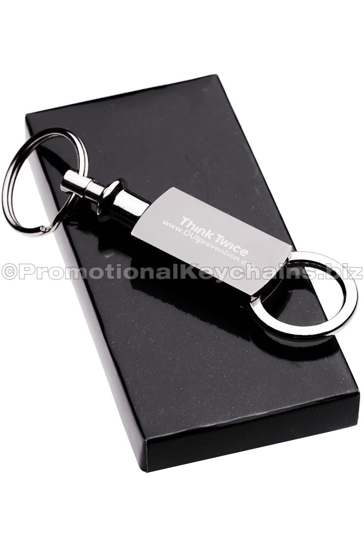 A polished metal keychain featuring a text engraving and a pull and twist attachment on the bottom. It is sitting on top of a black gift box.