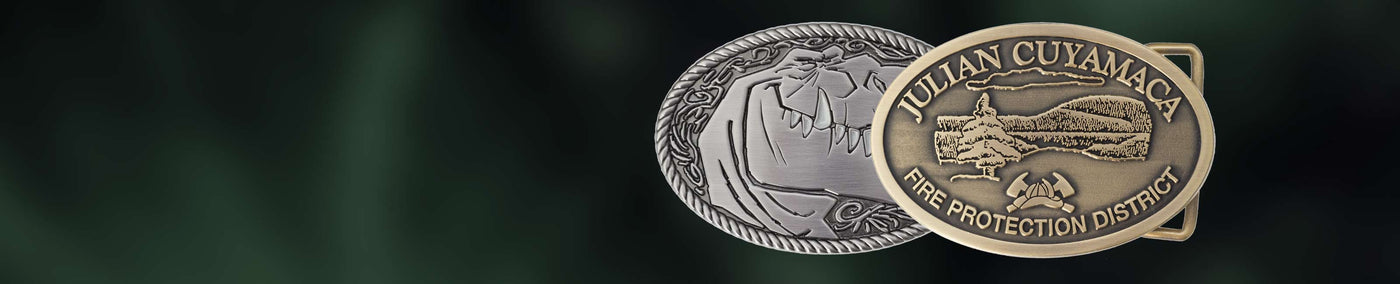 Two belt buckles, one nickel and one brass, with custom die cast designs on a dark background.