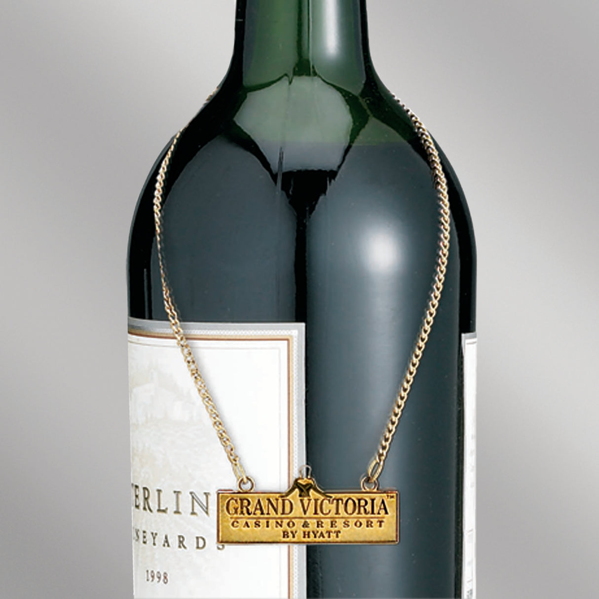 A green wine bottle adorned with a polished gold wine bottle charm on a golden chain, bearing the "Grand Victoria Casino & Resort by Hyatt" logo embossed on a rectangular tag.