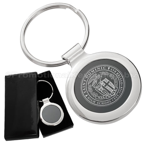 There are two images. On the left, a circular polished metal keychain featuring a black inlay displayed in a black gift box. On the right is the same keychain with a logo clearly engraved in the center.