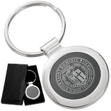 There are two images. On the left, a circular polished metal keychain featuring a black inlay displayed in a black gift box. On the right is the same keychain with a logo clearly engraved in the center.