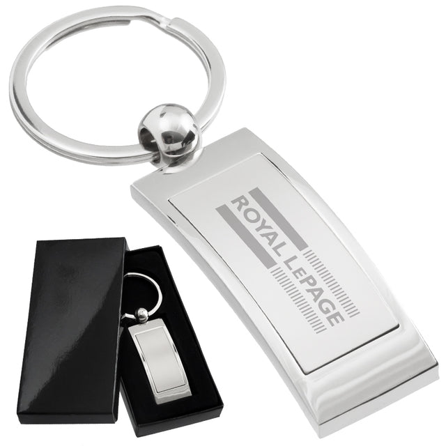 There are two images. On the left, a polished metal keychain, rectangular in shape, displayed in a black gift box. On the right is the same keychain with a logo clearly engraved in the center.