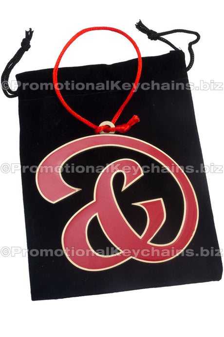 A custom shaped holiday ornament with a red and gold enamel design, hanging from a black bag with a red ribbon.